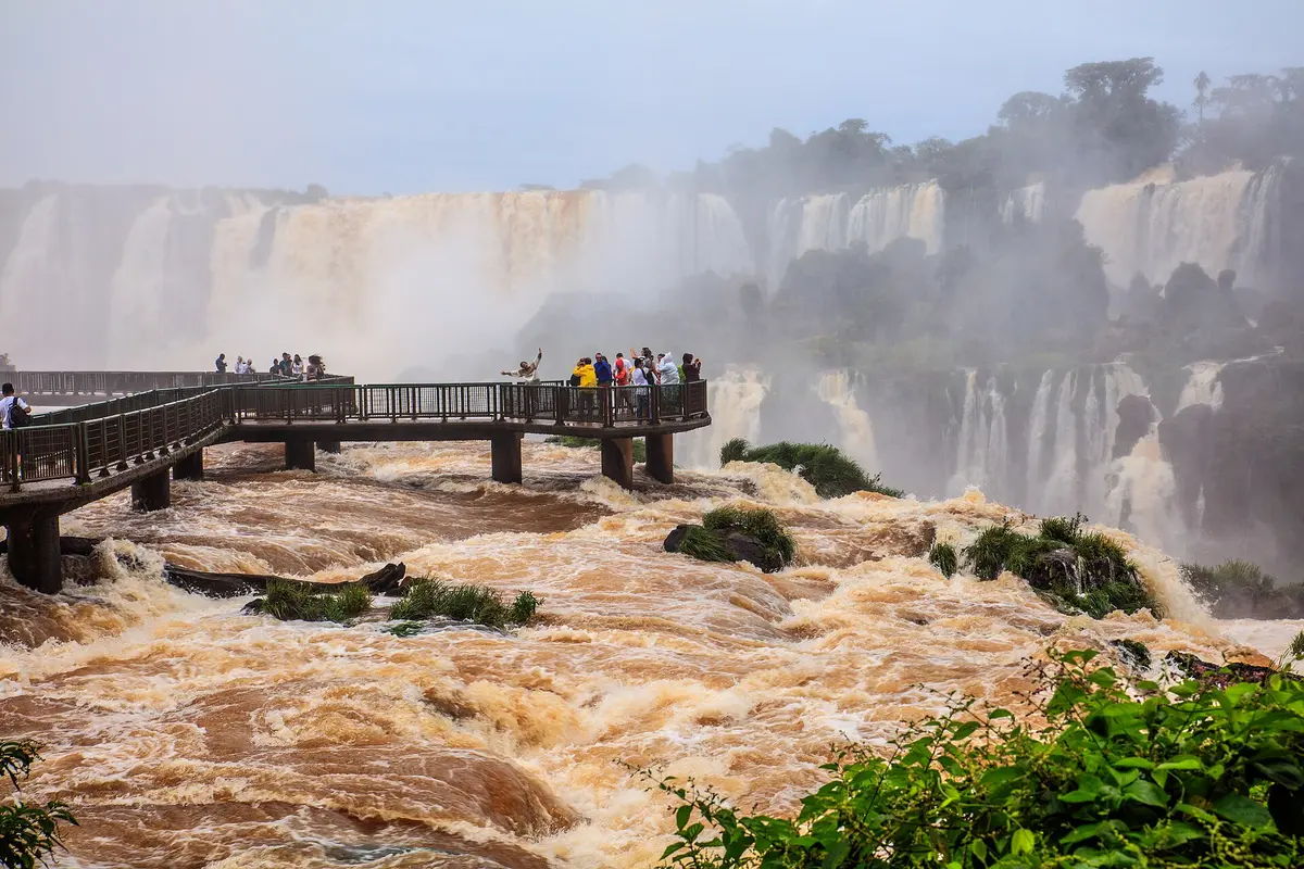 Visitors on the walkway over the brown, fast-flowing Iguazú River on the Brazilian side, with a long line of waterfalls behind in the mist