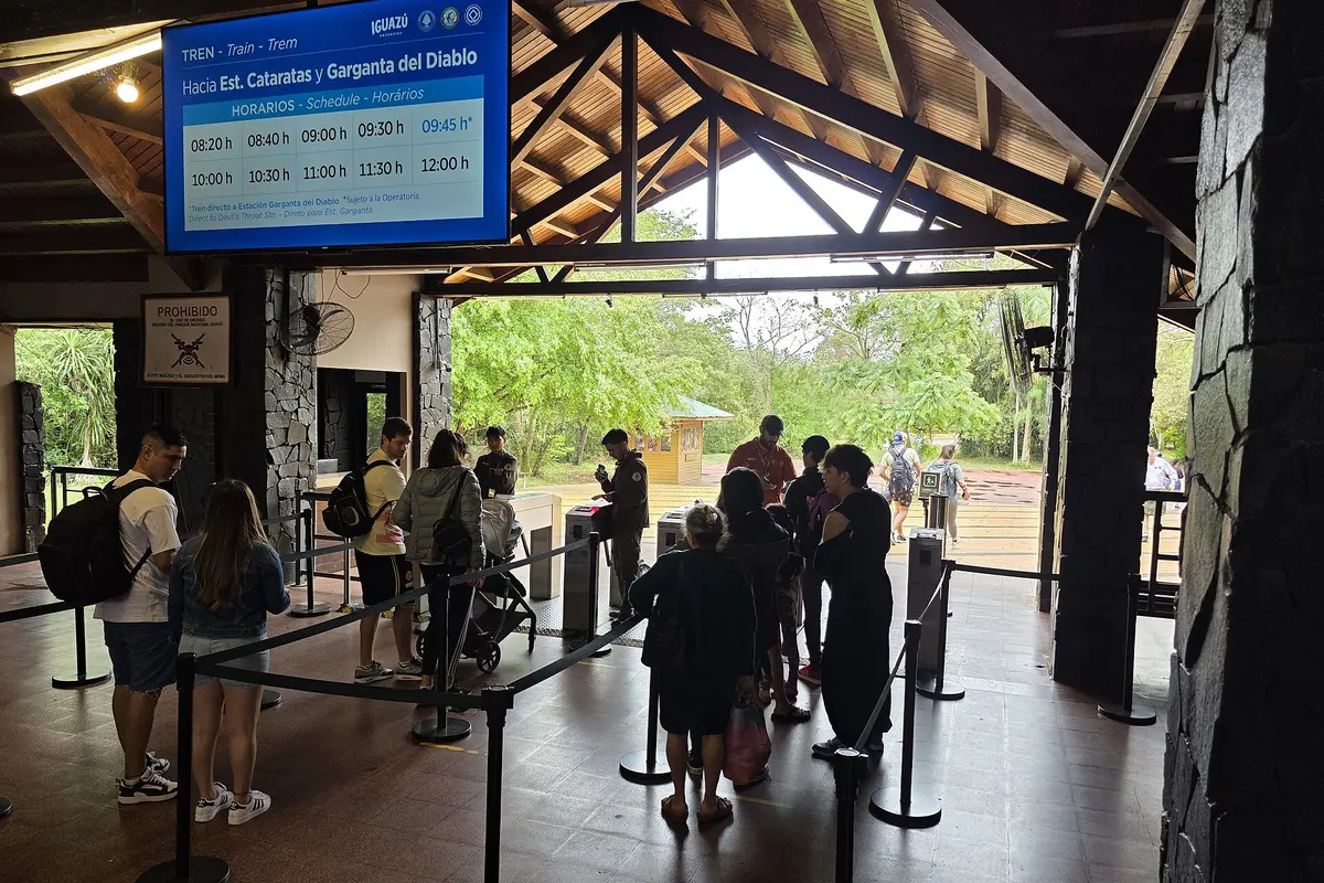 Visitors queueing at the gates for the Ecological Jungle Train in Iguazú National Park, under a screen showing departures to Cataratas and Garganta del Diablo stations